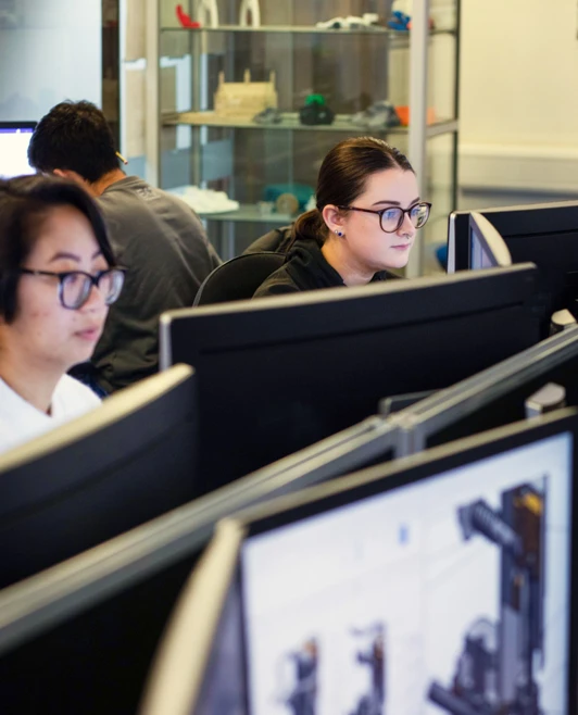 Students working on CAD designs at desktop computers in a classroom, with shelves displaying 3D-printed models in the background. Students working on CAD designs at desktop computers in a classroom, with shelves displaying 3D-printed models in the background.